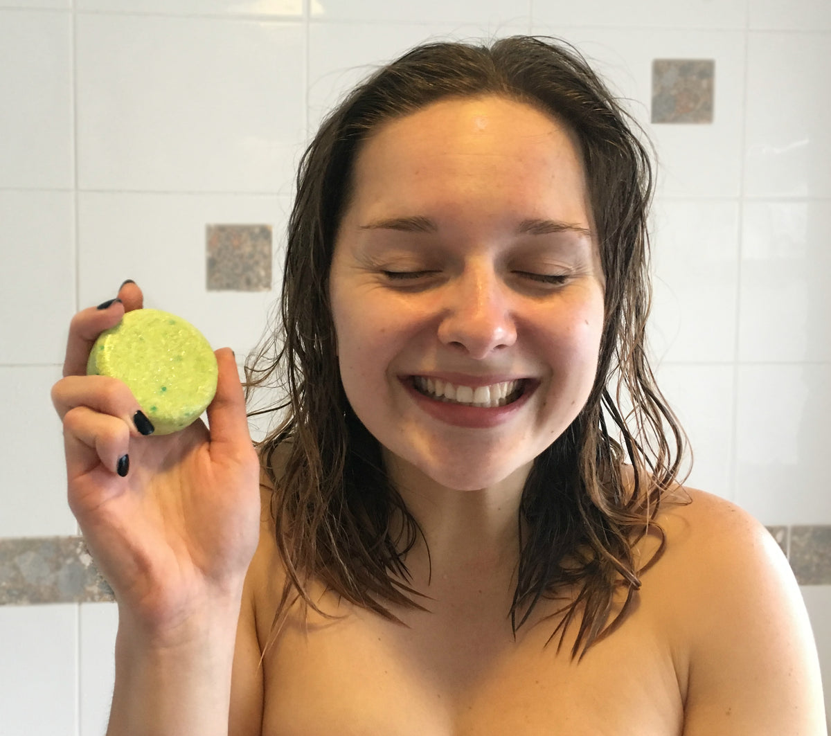 Set in bathroom shower. Picture of young woman smiling with her eyes closed, holding a shampoo bar in right hand.