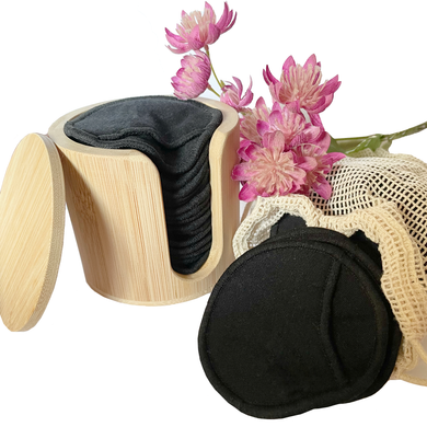 Bamboo cylindrical holder with 17 x black bamboo reusable make up remover pads, with more pads in a mesh laundry bag. Pink flowers adding some colour to the setting. 